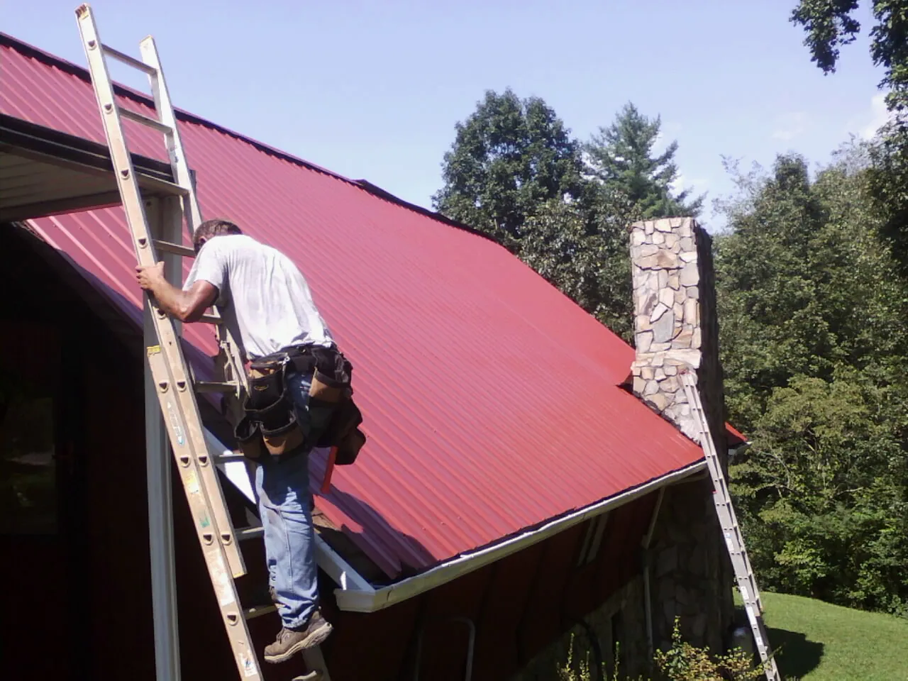 Roofer on a ladder beside a red metal roof and stone chimney — Soffit & Fascia Repair by Roofing Brothers in Depew